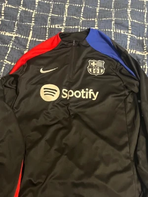 FC Barcelona träningsset Nike svart - Säljer ett svart FC Barcelona träningsset från Nike med klubbmärke och Spotify-logga på tröjan. Tröjan har dragkedja, röd och blå detalj på axlarna och lång ärm. Byxorna har klubbmärke och Nike-logga samt resår i midjan. Perfekt för fotbollsträning.