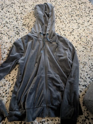 Mörkgrå velour hoodie med dragkedja stl xs - Mjuk och skön mörkgrå hoodie i velour med dragkedja framtill och stora fickor. Huva med breda svarta snören och ribbade muddar vid ärmslut och nederkant. Perfekt för en avslappnad och chill stil.