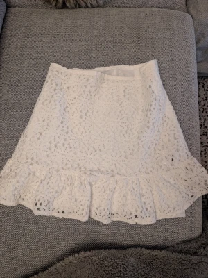 Vit spetskjol med volangkant stl xs - Supersöt vit kjol i spets med blommigt mönster och volangkant nertill. Kjolen är kort och har en feminin, romantisk vibe som passar perfekt till sommaren. Ingen synlig dragkedja eller knappar. Perfekt att styla med en enkel topp eller stickad tröja.