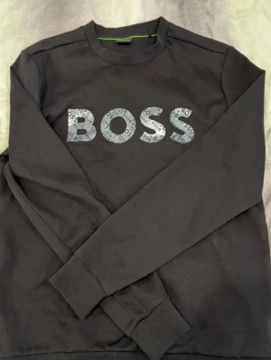 Svart tröja BOSS - Svart långärmad topp från BOSS med stor logga framtill i grått och mönstrad text. Klassisk rund halsringning och ribbade muddar vid ärmslut och nederkant. Helt ny, använd ett väldigt fåtal gånger.