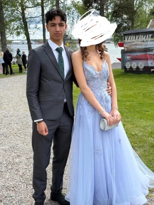 Ljusblå balklänning med spetsdetaljer - Långklänning i ljusblå ton med vacker spets och broderade detaljer. Klänningen har öppen rygg med snörning och tunna axelband som korsas. Fluffig kjol i flera lager och elegant v-ringad front. Perfekt för bal eller student.