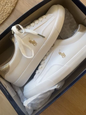 Vita och guldiga sneakers från Polo Ralph Lauren - Stilrena vita sneakers från Polo Ralph Lauren med guldfärgade detaljer och logga på sidan. Har aldrig används då de var förstora. Original pris 1000.