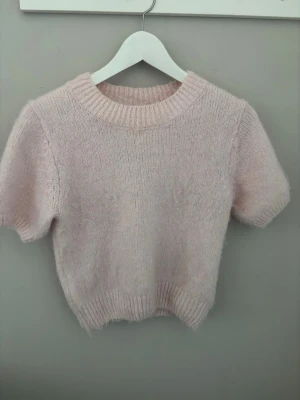 Ljusrosa stickad t-shirt - Supersöt ljusrosa stickad t-shirt med korta ärmar och ribbade muddar vid hals, ärmslut och nederkant. Tröjan har en mjuk och fluffig känsla, perfekt för att addera en gullig touch till din outfit.