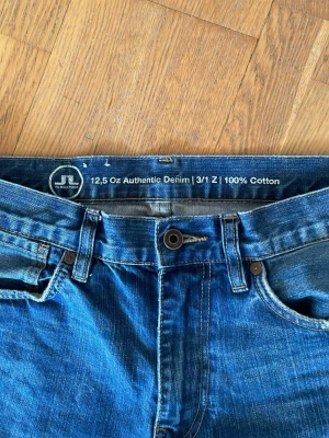 Blå jeans från J.Lindeberg - Säljer ett par klassiska blå jeans från J.Lindeberg i 100% bomull. Jeansen har fem fickor, normal midja och raka ben. Snygga kontrastsömmar och läderpatch med logga bak. Perfekta till en avslappnad stil.