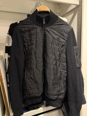 Svart cardiganjacka från J.Lindeberg - Snygg svart cardiganjacka från J.Lindeberg med quiltad front och stickade ärmar. Jackan har hög krage, dragkedja framtill och fickor med dragkedja. Ärmarna är ribbstickade och har en ficka på ena ärmen. Perfekt för lager på lager.