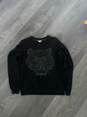 Svart sweatshirt från Kenzo med tiger - Cool svart sweatshirt från Kenzo med det ikoniska tigerhuvudet broderat framtill i grönt, blått och grått. Rund halsringning och ribbade muddar vid ärmslut och nederkant. Perfekt för dig som gillar streetwear och vill sticka ut.