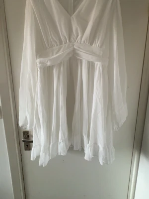 Vit omlottklänning med v-ringning - Säljer en vit kortklänning med v-ringning och omlottdetalj i midjan. Klänningen har långa, luftiga ärmar och ett flowy, lätt tyg som ger en romantisk känsla. Perfekt för sommarens alla tillfällen!