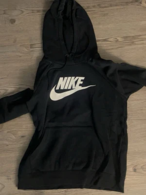 Svart hoodie Nike - Svart hoodie från Nike med stor vit logga framtill. Klassisk modell med huva och känguruficka. Mjuk och skön, passar till det allt.
