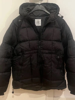 Svart pufferjacka med huva från Cheap Monday - Säljer en svart pufferjacka från Cheap Monday med huva och dragkedja framtill. Jackan har två sidofickor med dragkedja och justerbara snören i huvan och nederkanten. Quiltad design och oversized passform för en cool vinterlook.