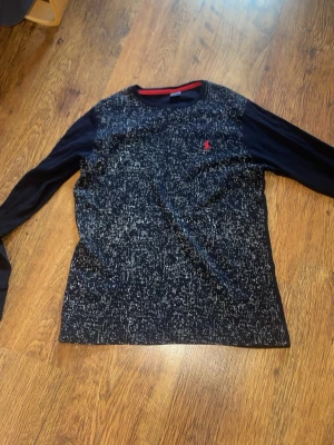 Svart långärmad tröja från Polo Ralph Lauren - Snygg svart långärmad tröja från Polo Ralph Lauren med vitt spräckligt mönster och klassisk röd logga broderad på bröstet. Tröjan har rund halsringning och är tillverkad i mjukt material som känns skönt mot huden. Perfekt för dig som gillar streetwear vibes. 100% äkta