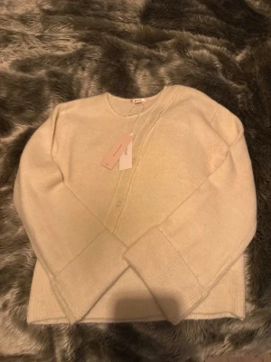 Krämvit kofta från Gina - Mysig krämvit kofta från gina i mjuk ullblandning. Koftan har rund halsringning, knäppning framtill och breda ribbade muddar vid ärmslut och nederkant. Enkel och stilren design som passar perfekt till lager-på-lager.