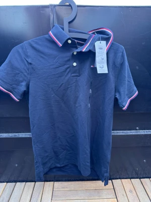 Svart pikétröja från Tommy Hilfiger🙌🤩 - Snygg marinblå pikétröja från Tommy Hilfiger med klassisk krage och knappar framtill. Tröjan har röda och vita detaljer på krage och ärmslut samt liten logga på bröstet. Tillverkad i bomull och har korta ärmar. Helt ny!🙌🤩