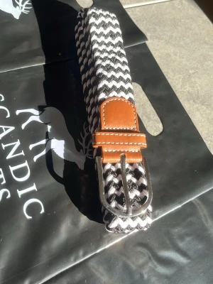 Svartvitt flätat bälte med läderdetalj - Snyggt flätat bälte i svart och vitt med bruna läderdetaljer vid spännet. Bältet har ett klassiskt silverfärgat metallspänne och passar perfekt till jeans eller chinos. Mönstret är randigt och ger en modern touch till din outfit. 105cm