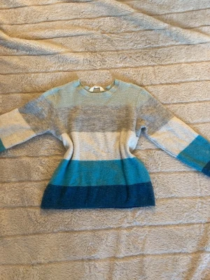 Randig stickad tröja från H&M - Mysig stickad tröja från H&M med breda horisontella ränder i blått, grått, beige och turkos. Tröjan har rund halsringning och långa ärmar. Perfekt för dig som gillar färg och vill ha en skön vardagströja.