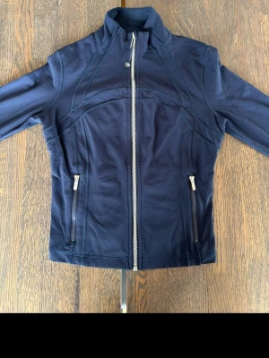 Marinblå träningsjacka från Lululemon - Snygg marinblå träningsjacka från Lululemon med figurnära passform, hög krage och hel dragkedja framtill. Två fickor med dragkedja på sidorna och diskret logga på ryggen. Tillverkad i stretchigt funktionsmaterial som passar perfekt till träning.