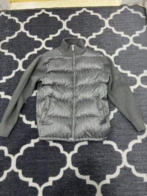 Grå dunväst med stickade ärmar - Snygg grå dunväst med diskret logomönster över hela västen. Västen har ribbstickade ärmar och hög krage i samma grå ton samt dragkedja framtill. Perfekt för dig som vill ha en exklusiv och trendig look.