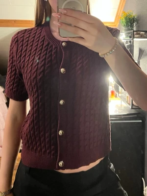 Vinröd kabelstickad kofta kort ärm - Snygg vinröd kabelstickad kofta med korta ärmar och guldfärgade knappar framtill. Rund halsringning och ribbade muddar vid ärmslut och nederkant. Knappt använd, storlek L men passar mig som har S i vanligq fall. 
