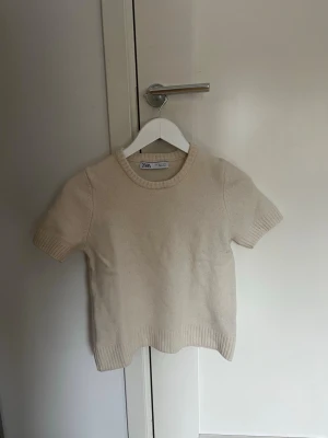 Beige stickad topp från Zara - En enkel och stilren beige stickad topp från Zara med korta ärmar och rund halsringning. Perfekt att matcha med jeans eller kjol för en clean look. Mjuk och skön kvalitet.