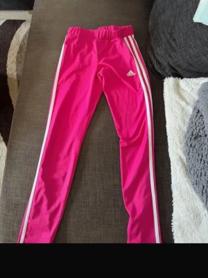 Rosa träningsbyxor från Adidas - Säljer ett par snygga rosa träningsbyxor från Adidas med vita ränder längs sidorna och Adidas-logga på låret. Byxorna har en smal passform och resår i midjan. Perfekta för gymmet eller löprundan. Materialet känns mjukt och stretchigt.
