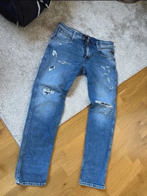 Blå slitna jeans Replay Anbass - Säljer ett par blå jeans från Replay, modell Anbass, med slim fit passform. Jeansen har slitningar och hål för en trendig, avslappnad look. Klassisk femficksmodell med dragkedja och knapp. Perfekta för dig som gillar streetstyle.