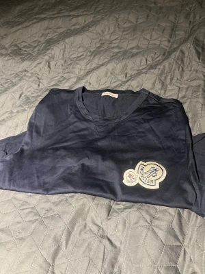 Mörkblå Moncler t-shirt med patch - Snygg mörkblå t-shirt från Moncler med klassisk rund hals och en stor broderad patch på bröstet. Tillverkad i mjuk bomull som känns skön mot huden. Perfekt för dig som gillar stilrena och exklusiva plagg.