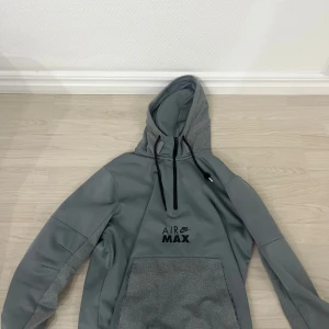 Grå Nike Air Max hoodie med huva - Säljer en grå hoodie från Nike Air Max med stor magficka och justerbar huva med snörning. Hoodien har tryckt logga på bröstet och är tillverkad i mjukt material, perfekt för en avslappnad streetwear-look.