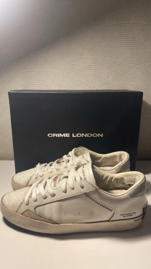 Vita Crime London sneakers - Säljer ett par vita sneakers från Crime London med coola detaljer som svart söm och text på sidan. Skorna har lågt skaft, vita snören och en klassisk siluett. Materialet är läder med inslag av mocka vid tån. Perfekta för dig som gillar streetstyle och enkel design.