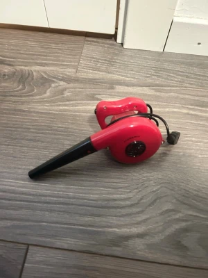 Röd elektrisk blåsmaskin - Elektrisk blåsmaskin i röd plast med svart munstycke och handtag. Perfekt för att blåsa bort damm eller smuts från svåråtkomliga platser. Utrustad med strömkabel och kompakt design som gör den enkel att använda och förvara.