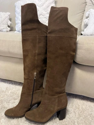 Bruna höga mockastövlar med klack - Säljer ett par bruna overknee-stövlar i mjuk mocka med chunky klack och dragkedja på insidan. Stövlarna har en stilren design och passar perfekt till höst- och vinterlooken. De är höga och går över knät för en trendig vibe.