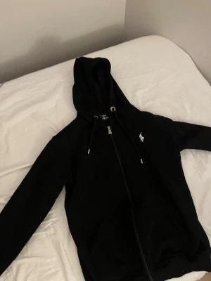 Svart hoodie från Ralph Lauren - Säljer en svart hoodie från Ralph Lauren med dragkedja framtill och klassisk vit logga på bröstet. Hoodien har justerbar huva med snören och ribbade muddar vid ärmar och midja. Perfekt för en chill och stilren look.