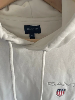 Vit hoodie från GANT S/M - Säljer en stilren vit hoodie från GANT med broderad logga och sköld på bröstet. Storleken är M med mer som S.