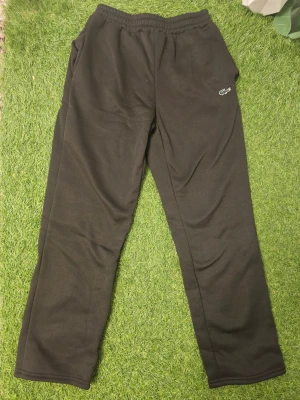 Svarta mjukisbyxor från Lacoste - Säljer ett par svarta mjukisbyxor från Lacoste med klassisk krokodillogga på ena benet. Byxorna har resår i midjan och raka ben. Perfekta för chill dagar eller när du vill ha det bekvämt. Materialet känns mjukt och skönt mot huden.