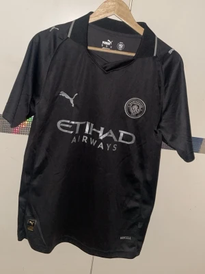 Svart Manchester City matchtröja Puma - Snygg svart Manchester City matchtröja från Puma med klubbmärke och Etihad Airways-tryck på bröstet. Tröjan har korta ärmar, sportig passform och är tillverkad i lätt och ventilerande material med DryCell-teknologi.