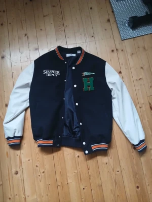 Stranger Things bomberjacka svart/vit - Snygg svart bomberjacka med vita ärmar och broderade detaljer från Stranger Things. Jackan har orange och vita ränder på muddar och krage, samt coola patches med text och bokstaven H på bröstet. Perfekt för dig som gillar retrostil och populärkultur. HM, stl 152. Men basiccly storlek S. Jag är 173cm o den passar mig. Inte använd