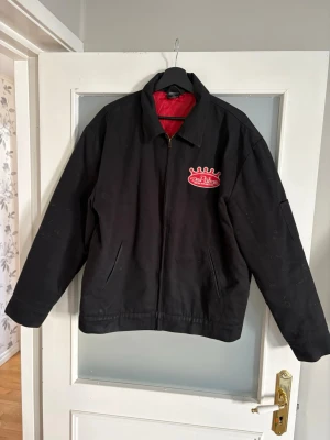 Svart Depalma jacka med streetmotiv storlek M - Svart jacka från Depalma med dragkedja och röd insida. På bröstet finns en broderad logga och på ryggen ett stort streetmotiv med texten 'Street Slayer' och en röd hot rod-bil. Jackan har klassisk krage och sidofickor. Perfekt för dig som gillar streetwear och bilar.