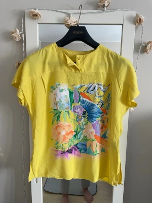 Gul tuzzi  blus med färgglatt tryck - Snygg Tuzzi gul blus med korta ärmar och färgglatt blommigt tryck framtill. Blusen har en liten knappdetalj vid halsen och ett mindre tryck på ryggen. Perfekt för dig som gillar färg och vill sticka ut lite extra. Materialet känns lätt och luftigt.vet inte vad det är för material,tror det är bomull .