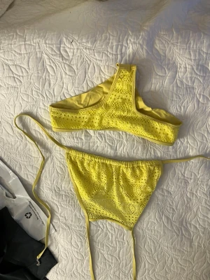 Gul bikini med spetsdetaljer - Snygg gul bikini med spetsdetaljer och trendig design. Överdelen har hög halsringning och underdelen har smala knytband i sidorna. Perfekt för soliga dagar på stranden eller vid poolen.