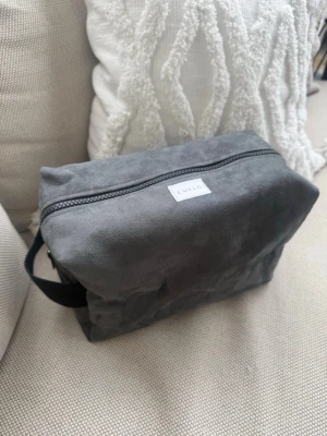 Grå necessär från Emelo i alcantara mockaimitation - Snygg och stilren grå necessär från Emelo i mjuk alcantara mockaimitation. Väskan har en rektangulär form, dragkedja upptill och ett praktiskt handtag på sidan. Svart insida med innerfack. Perfekt för att organisera dina småsaker eller smink när du är på språng. Storlek large. Mått ca 27x17x12cm