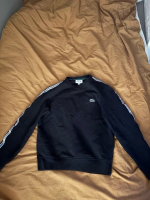 Svart sweatshirt från Lacoste - Snygg svart sweatshirt från Lacoste med klassisk krokodillogo på bröstet och vita logoband längs ärmarna. Tröjan har rund halsringning och ribbade muddar vid ärmslut och nederkant. Perfekt för en sportig och stilren look. 