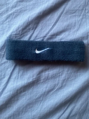 Svart Nike pannband i frotté - Säljer ett svart pannband från Nike med den klassiska vita swoosh-loggan broderad framtill. Tillverkat i mjuk och stretchig frotté som håller håret på plats och absorberar svett. Perfekt accessoar för sportiga looks eller när du vill hålla håret borta från ansiktet.kan diskutera pris
