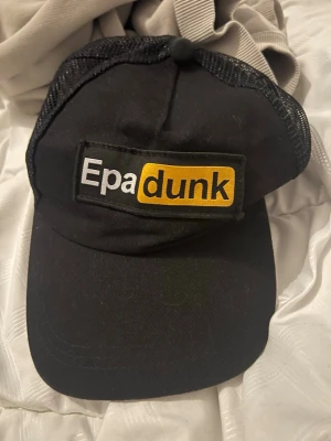 Svart truckerkeps med Epadunk-tryck - Säljer en svart truckerkeps med mesh på sidorna och ett stort broderat märke framtill med texten 'Epadunk' i vitt och gult. Kepsen har böjd skärm och justerbar snapback baktill. Perfekt för dig som gillar streetstyle och vill sticka ut.