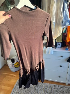 Brun ribbad kortklänning - Säljer en brun ribbad kortklänning med trekvartsärm och volang nedtill. Klänningen har en enkel, stilren design och är tillverkad i ett mjukt material som sitter bekvämt på kroppen. Perfekt för dig som gillar en avslappnad men trendig look.