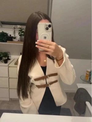 Beige kavaj med bruna spännen - Stilren beige kavaj med två bruna spännen framtill som stängning. Kavajen har en rak passform och är utan krage, vilket ger en modern look. Perfekt att bära över en topp eller klänning för en trendig outfit.