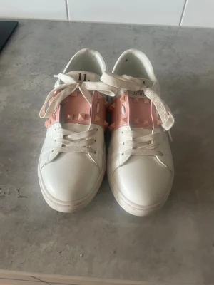 Valentino Rockstud Untitled sneakers - Snygga vita sneakers från Valentino med rosa detaljer och ikoniska Rockstud-nitar. Skorna har vita snören och en låg silhuett. Tillverkade i läder med coola rosa paneler och nitar på sidorna. Perfekta för dig som vill sticka ut med din stil.