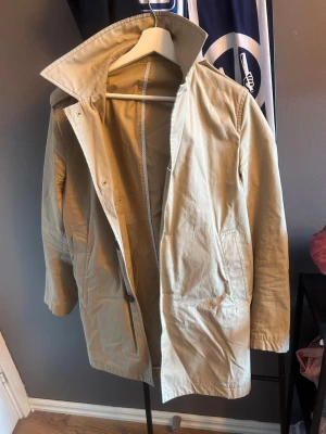 Beige trenchcoat med hög krage - Stilren beige trenchcoat med hög krage och knäppning framtill. Jackan har två sidofickor och är tillverkad i ett lätt bomullsliknande material. Perfekt för lager-på-lager och en clean look. Passar dig som gillar klassiska och minimalistiska plagg.