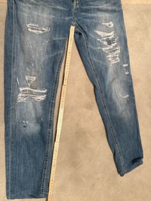 Blå slitna jeans från dondup - Säljer ett par blå jeans från dondup med tydliga slitningar och trasiga detaljer på både fram- och baksida. Jeansen har klassisk femficksmodell och normal midja. Materialet är denim i bomull och passformen är rak. Perfekta för en avslappnad och trendig look. små.