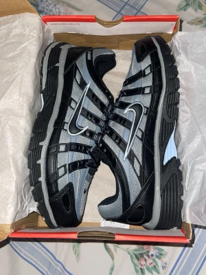 Nike P-6000 svart och grå sneakers - Nike P-6000 sneakers i svart och grått med vit swoosh på sidan. Skorna har en sportig design med mesh och syntetiska paneler för bra andningsförmåga och komfort. Tjock sula i svart och grått ger en chunky look. Perfekta för dig som gillar retroinspirerad stil.