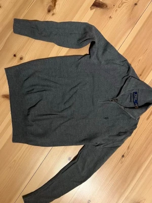 Mörkgrå stickad tröja Polo Ralph Lauren - Snygg mörkgrå stickad tröja från Polo Ralph Lauren med halv dragkedja vid halsen och diskret broderad logga på bröstet. Tröjan har lång ärm och smal passform, perfekt för lager på lager under kyliga dagar. Står XS på tröjan men fittar en S.