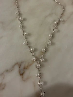 Silverfärgat halsband med pärlor - Elegant halsband i silverfärg med vita pärlor längs kedjan. Halsbandet har en Y-formad design där pärlorna är jämnt placerade och ger ett stilrent och klassiskt intryck. Perfekt accessoar för att lyfta din outfit med en touch av elegans.
