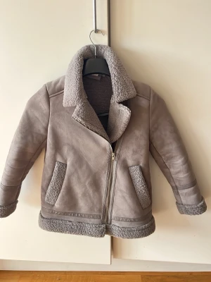 Grå teddyjacka från Zara Girls - Supersnygg grå teddyjacka från Zara Girls med mjukt foder och detaljer i teddy på krage, fickor och ärmslut. Jackan har dragkedja framtill och två fickor. Perfekt för kalla dagar och ger en trendig look.